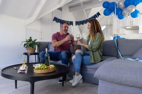Middle-aged couple toasting wine on grey sofa celebrating intimate birthday with balloons