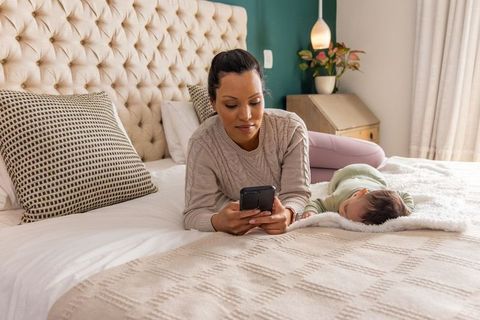 Mother using smartphone while baby naps on cozy bed, modern home bonding moment
