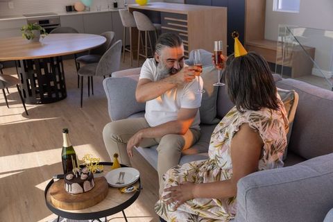 Middle-aged couple clinking champagne flutes on sofa celebrating intimate home birthday