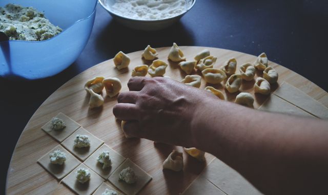 This image captures a person handcrafting tortellini pasta on a wooden cutting board, with dough squares filled with ricotta cheese. Ideal for depicting culinary arts, traditional Italian cooking, or DIY cooking activities. Useful in cookbooks, cooking blogs, tutorials, and gourmet magazines.