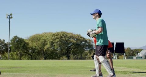 Wicketkeeper standing ready on sunny cricket field wearing teal shirt gloves pads and cap