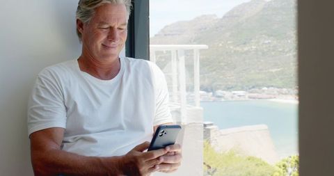 Senior man relaxing with smartphone on sunlit seaside balcony overlooking bay and coast