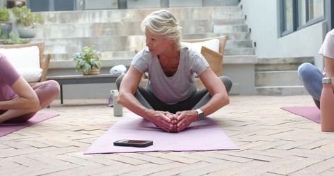 Mature woman stretching during outdoor yoga class on brick patio with pink mat and smartphone