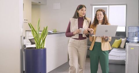 Diverse female coworkers walking office lounge holding mug and checking laptop screen