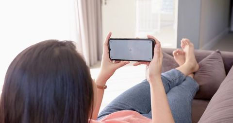 Mid-adult asian woman reclining on sofa watching smartphone in minimalist living room