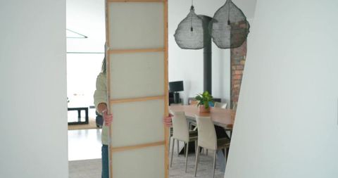 Mature woman holding wooden canvas panel while moving artwork through modern dining room