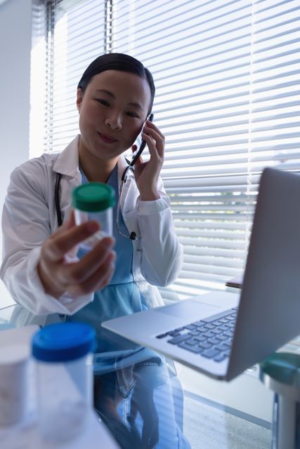 Female doctor holding a bottle of pills while talking on a mobile phone in a clinic. Ideal for use in healthcare, medical consultation, telemedicine, and patient care contexts. Useful for illustrating modern healthcare practices, doctor-patient communication, and the use of technology in medicine.