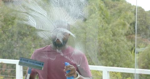 African man cleaning balcony glass with squeegee and spray bottle wearing maroon shirt