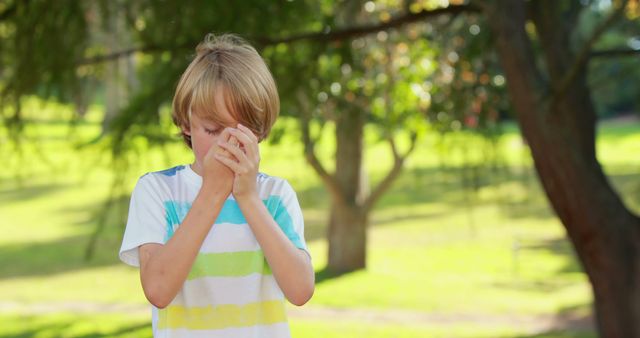 Young boy with blonde hair sneezing in a park during springtime, highlighting seasonal allergies. Scene emphasizes outdoor activities in nature and health considerations related to allergies. Ideal for use in articles or campaigns discussing children's health, seasonal allergies, benefits of outdoor activities, or healthcare essentials for kids.
