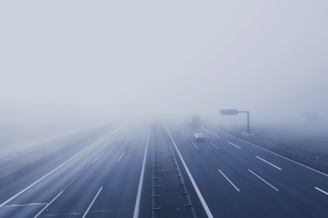 Morning highway with minimal traffic, veiled in thick fog. Two vehicles drive into the distance, creating a sense of mystery and calm. Perfect for themes like transportation challenges, driving conditions, safety concerns, or atmospheric elements in travel.