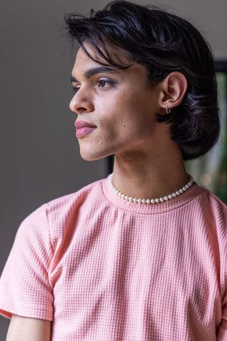 Man gazing out window wearing pearl necklace and pink textured tee, reflecting side profile