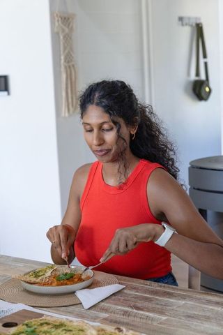 African american woman in red top wearing smartwatch enjoying curry with naan at sunlit dining table