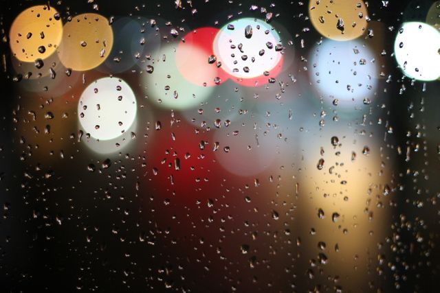 Raindrops on window with colorful blurred city lights in the background, creating a bokeh effect. Visual representation of a rainy night in the city. Ideal for users looking to illustrate themes of urban life, melancholy, romance, or reflection. Suitable for use in blogs, websites, advertising, or design projects needing an atmospheric and moody element.