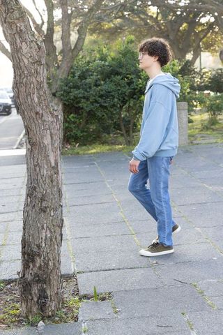 Teenager walking on tree-lined sidewalk wearing hoodie, jeans and glasses urban lifestyle