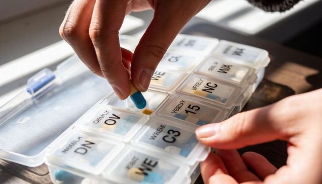 Hands placing blue-yellow capsule into labeled weekly pill organizer medication management