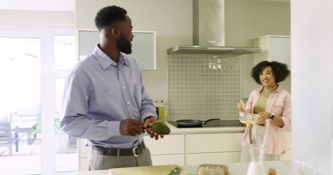Mid-adult couple preparing sunlit breakfast in kitchen, slicing avocado and whisking eggs