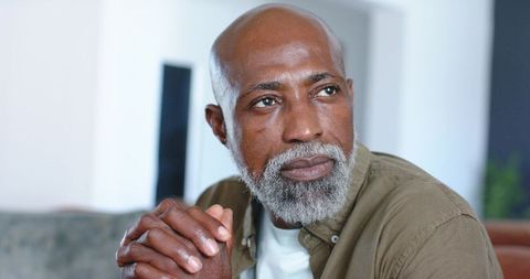 Thoughtful mature man sitting on sofa clasping hands gazing sideways for calm lifestyle
