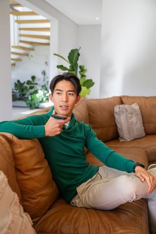 Asian man relaxing on leather sofa holding voice remote in modern minimalist living room