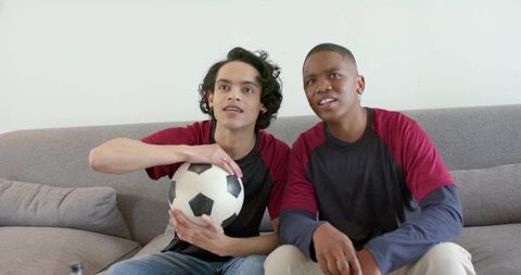 Diverse male friends watching soccer match on couch holding ball cheering for team