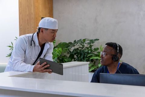 Asian doctor consulting black receptionist at clinic desk showing tablet and discussing care