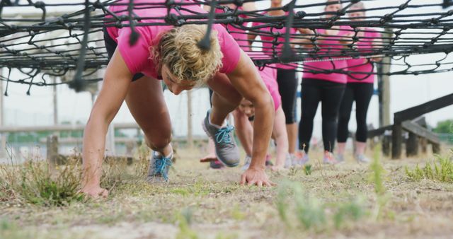 Caucasian female friends in pink t shirts watching and crawling under nets at bootcamp. Female fitness, friendship, challenge and healthy lifestyle.
