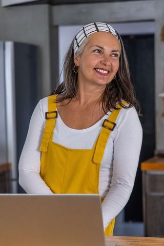 Smiling mature woman wearing yellow overalls using laptop in modern cozy kitchen