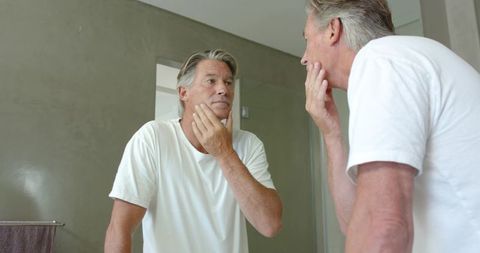 Middle-aged man checking face in bathroom mirror touching chin during morning grooming routine