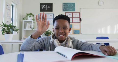 Preteen african american boy raising hand in bright classroom with headphones and notebook