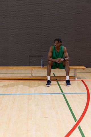 African american basketball player sitting on bench in gym wearing green jersey