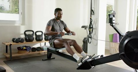 African american man rowing on ergometer in bright home gym with kettlebells and dumbbells