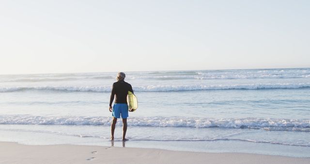 Surfer holding surfboard and looking at the ocean on a tranquil beach at sunrise. Ideal for themes related to relaxation, adventure, water sports, and summer vacations. Perfect for travel brochures, sports advertisements, and lifestyle blogs.