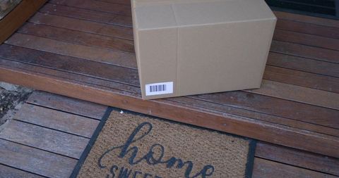 Sealed cardboard parcel sitting on porch step near welcome doormat on wooden decking