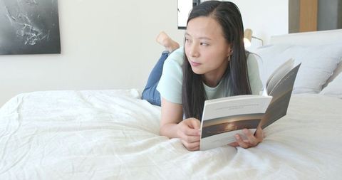 Asian woman lying on bed reading paperback in serene minimal bedroom with natural light