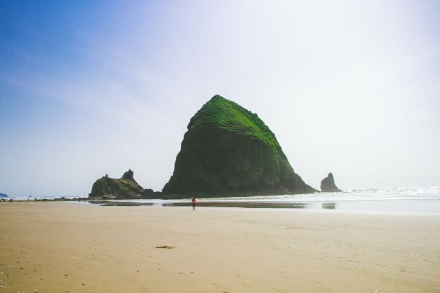 A person is seen walking alone on a vast sandy beach near a prominent rock formation during daytime. This scene displays a serene and peaceful seascape emphasizing the natural beauty of the coastline. Perfect for use in travel blogs, nature retreats, meditation platforms, or coastal tourism promotions.
