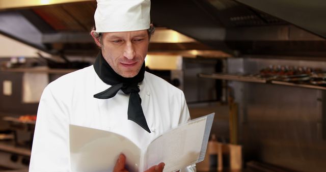 Chef standing in restaurant kitchen holding and reviewing recipe. Wearing standard chef uniform with hat and black necktie. Scene suggests culinary precision, professional kitchen environment, and meticulous preparation. Useful for illustrating professional cooking, restaurant operations, and chef roles.