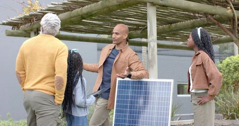 Technician explaining solar installation to diverse family under pergola, child holding tablet