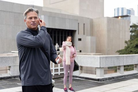 Middle-aged man adjusting wireless earbud on rooftop while woman prepares for workout
