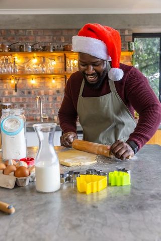 African American man wearing Santa hat rolling cookie dough in cozy rustic kitchen