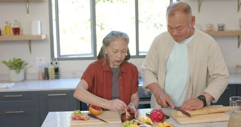 Senior asian couple preparing fresh fruit and slicing baguette together in bright kitchen