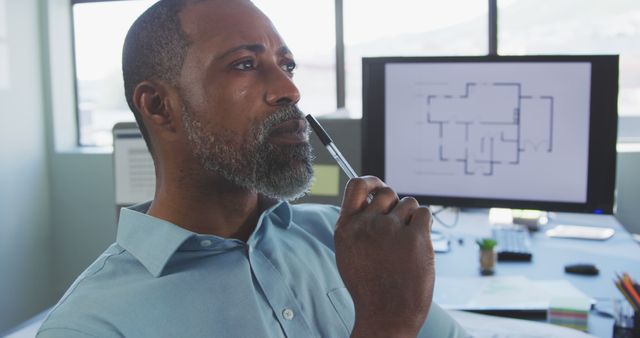 Mature African American architect contemplating design ideas at workstation with blueprints on computer screen. Suitable for use in articles or promotional material on architecture, creative professions, office environments, and professional occupations.