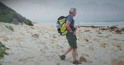 Senior Hiker Trekking along Rocky Beach with Backpack and Poles