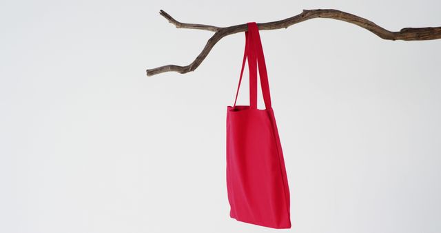 A bright red tote bag hangs from a bare tree branch, with copy space. Its vibrant color stands out against the minimalist background, suggesting themes of nature and sustainability.