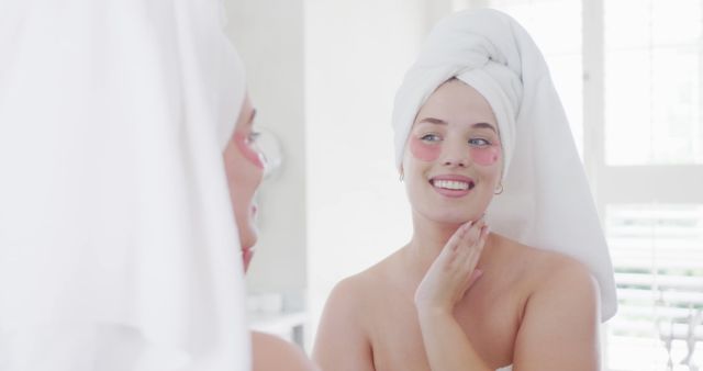Happy plus size caucasian woman wearing towel applying under eye beauty mask in sunny bathroom. Beauty, self care, wellbeing and healthy lifestyle, unaltered.