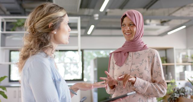 Two female professionals are shown engaging in a positive office discussion. One woman is wearing a hijab, emphasizing workplace diversity and inclusion. The setting is a modern office with lots of natural light and plants, indicating a forward-thinking, aesthetically pleasing work environment. This image is ideal for use in articles or social media posts related to teamwork, business communication, diversity in the workplace, or women in business.