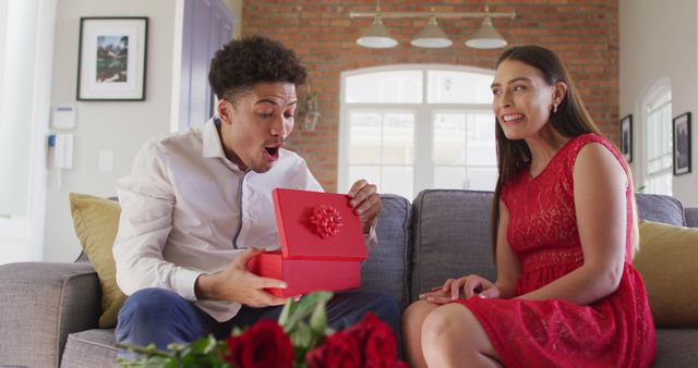 Man and woman sitting on sofa sharing gift. He has a surprised expression while looking at a red gift box. Ideal for use in advertisements for celebrations, romantic gifts, and lifestyle blogs.