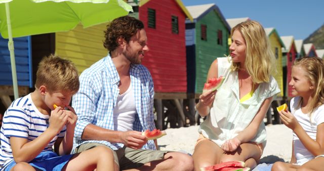 A Caucasian family enjoys a sunny beach day, eating watermelon with colorful beach huts in the background. Their relaxed posture and smiles convey a sense of leisure and family bonding time.
