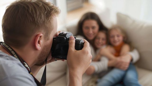 Male capturing mother and daughters cuddling on sofa with dslr in bright living room