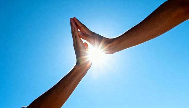 Raising forearms meeting palm-to-palm against bright blue sky with sunburst flare