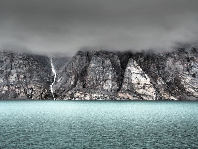 Dramatic cliff faces descending into turquoise glacier water, shrouded in mist. Perfect for use in tourism promotions, travel blogs, nature documentaries, geological studies, and environmental conservation materials, emphasizing the rugged beauty and untamed nature of remote landscapes.