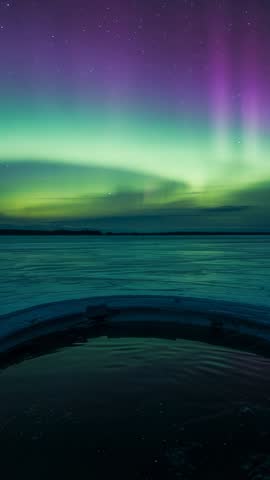 Vertical video showing northern lights reflecting in round ice hole, rippling frozen lake